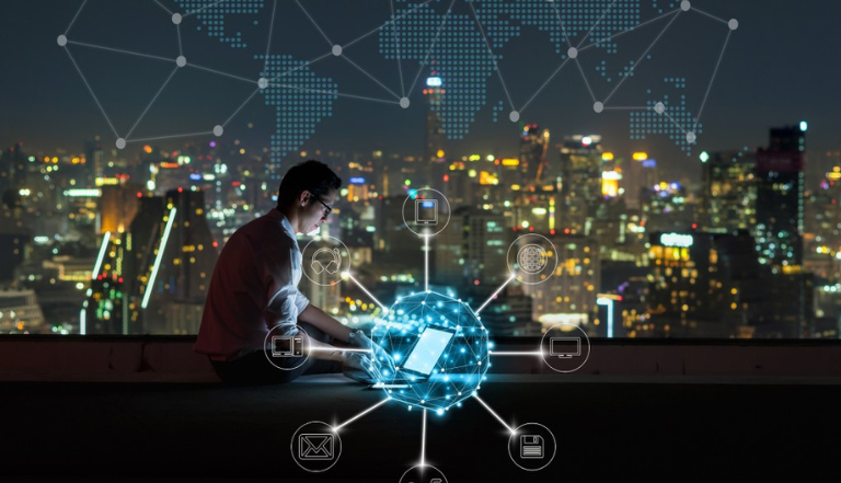 Man sits on roof of high building at night, looking down at a latptop, with glowing lights of city skyscapers in the background. digital tech images surround the laptop in a circle and emit a glowing network around the laptop.
