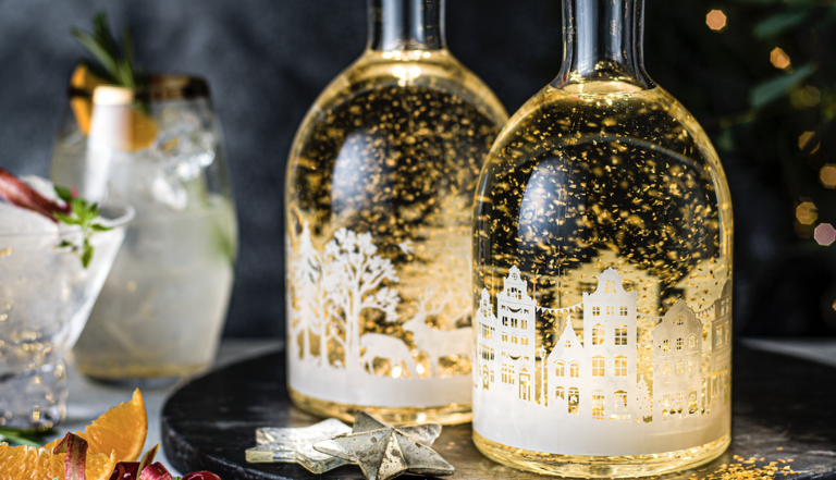 two festive gin bottles glowing with gold pieces floating inside, sitting alongside two out of focus festive cocktails