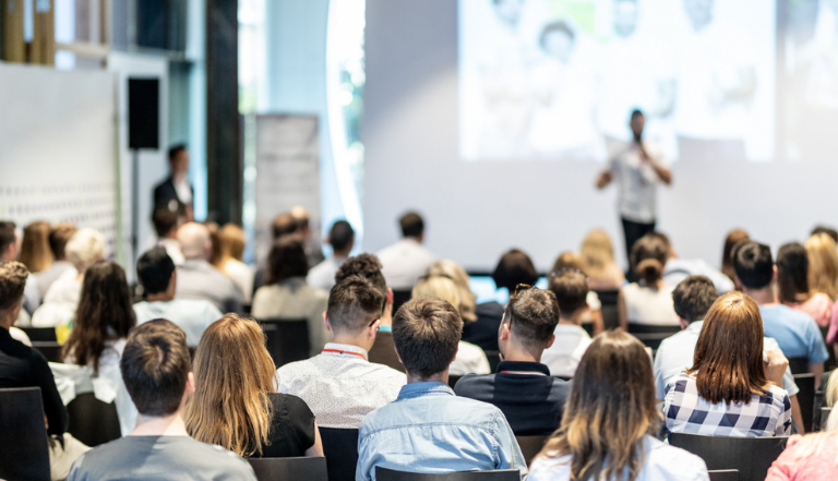 audience watching a presentation, with the speaker and presentation screen blurred in the background