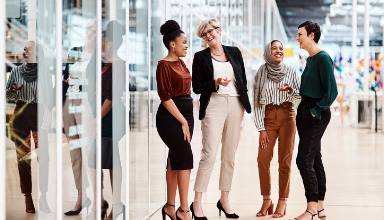 a group of 4 business women stand outside a modern office, chatting and laughing in front of glass panelling, with an office setting blurred in the background.
