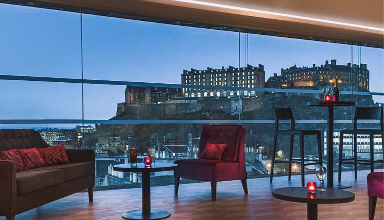 modern restaurant set up in low-lighting, soft red chairs and tables with candles overlooking a view of Edinburgh Castle at dusk through glass panels in the background